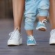 De eerste stappen van het kind, mamma in witte sneakers met een jonge zoon, gekleed in blauwe broek en blauwe schoenen, een warme zomerdag en leren lopen in de straat, de eerste stappen, de moeder houdt haar zoon.